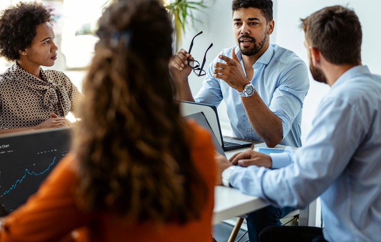 A group of colleagues sat around a table with their computers having a deep discussion