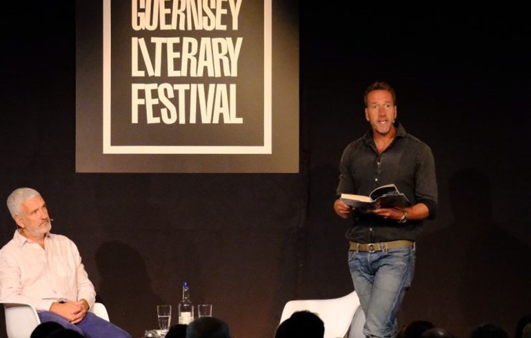 A man with white hair sitting down on stage listening to another gentleman reading a section from a book at the Guernsey Literary Festival