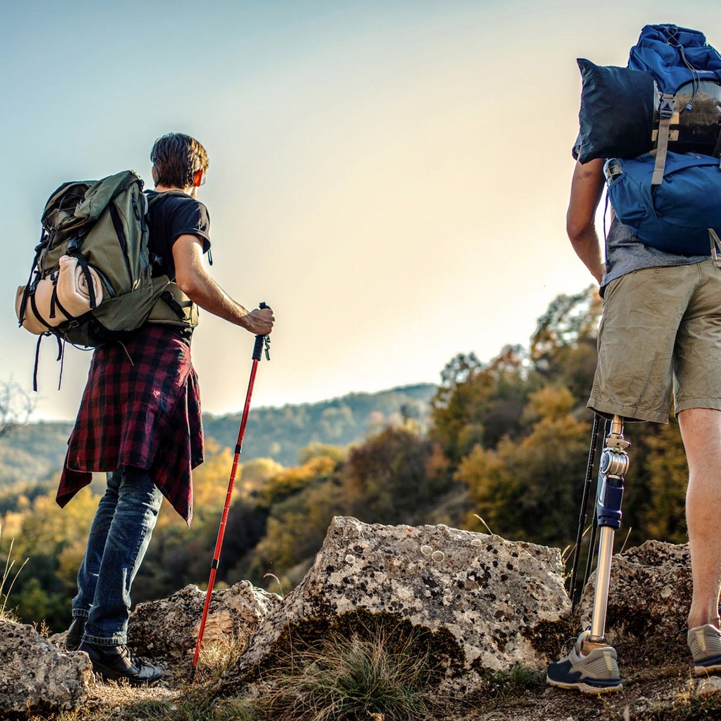 2 people hiking in the mountains with heavy backpacks on their backs. They are looking out into the distance. One of the hikers is amputee