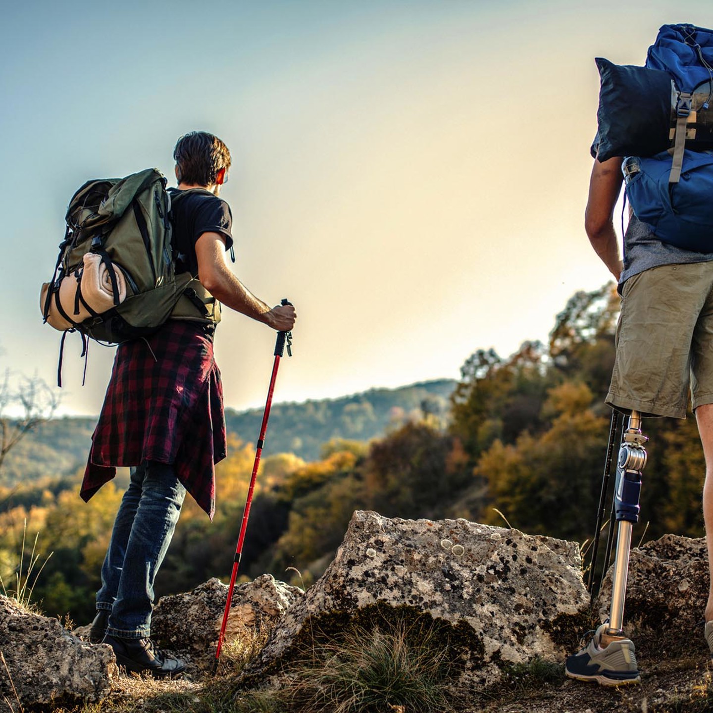 2 people hiking in the mountains with heavy backpacks on their backs. They are looking out into the distance. One of the hikers is amputee