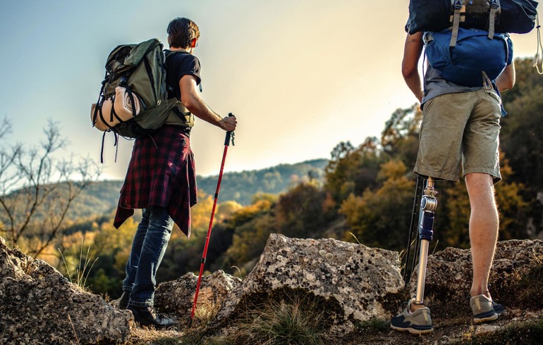2 people hiking in the mountains with heavy backpacks on their backs. They are looking out into the distance. One of the hikers is amputee