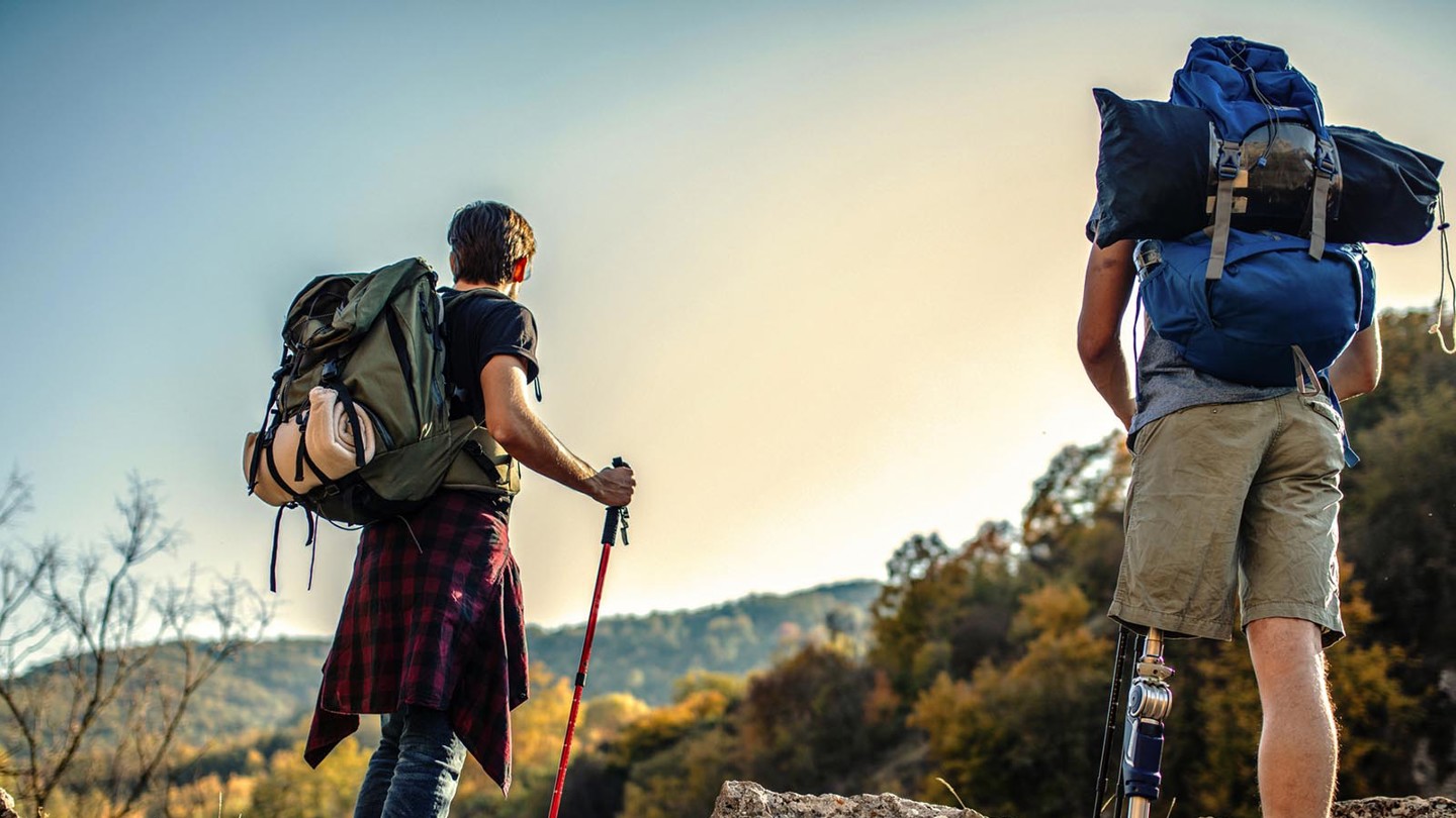 2 people hiking in the mountains with heavy backpacks on their backs. They are looking out into the distance. One of the hikers is amputee