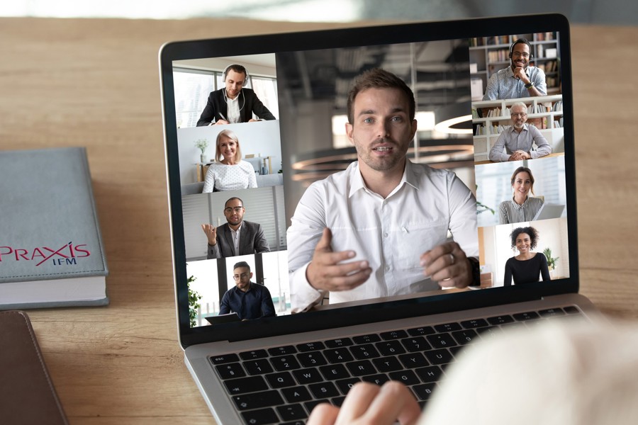 A laptop on a wooden table displaying a group of colleagues on a business video call. There is a Praxis IFM notebook also on the table.