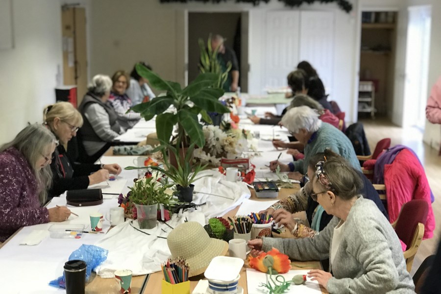 A group of elderly woman gathered around a long table getting creative drawing and painting the plants that are located in the middle of the table.