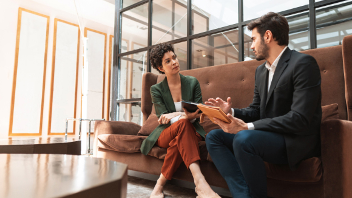 Two colleagues sat in a lobby having a discussion. The man is holding a hand held device while speaking to the woman