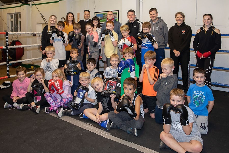 Many children sat in a boxing ring, wearing boxing gloves, smiling for a group photo