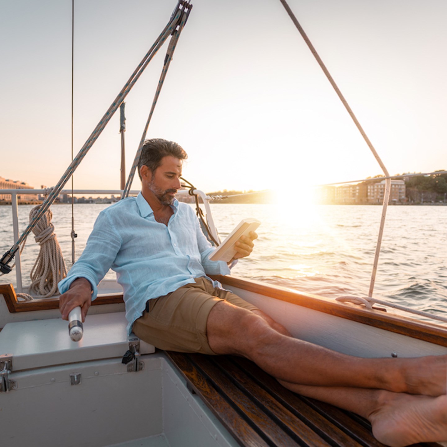 A man reading a book on a sail boat as the sun is setting