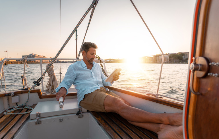 A man reading a book on a sail boat as the sun is setting