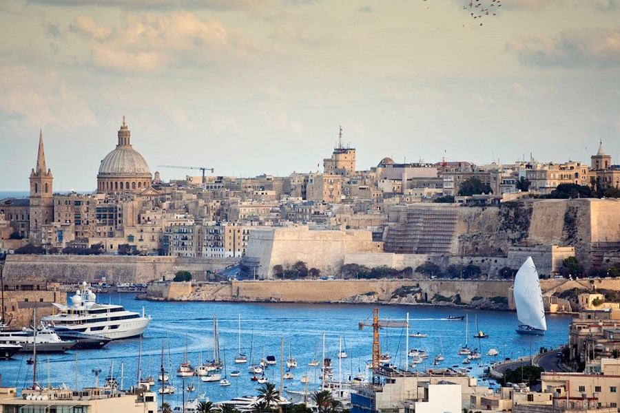 A river with many boats docked. A view of Malta architecture in the background