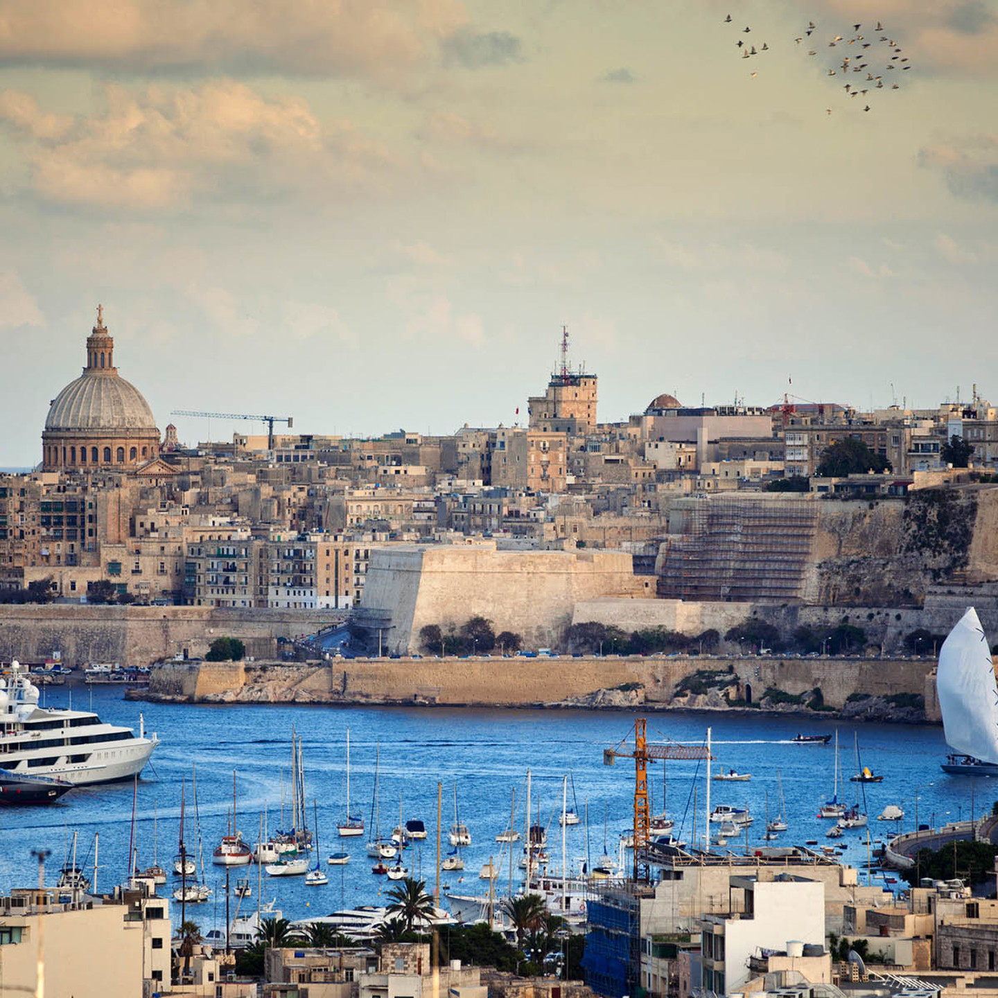 A river with many boats docked. A view of Malta architecture in the background