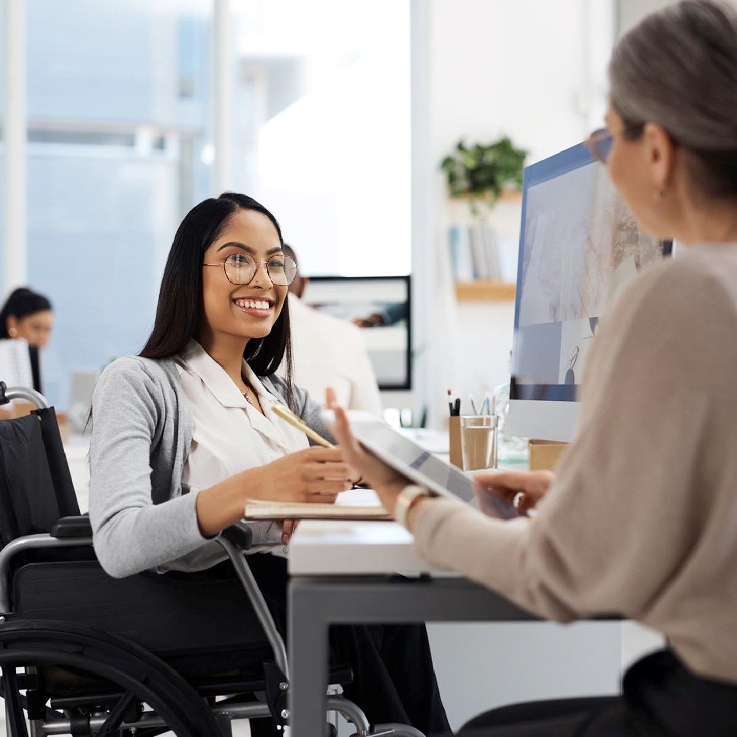 A woman in a wheelchair having a pleasant conversation with another woman while sat at her desk