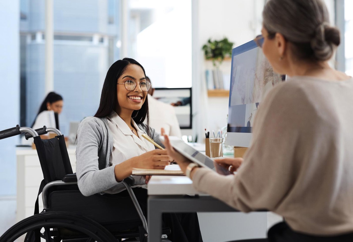 A woman in a wheelchair having a pleasant conversation with another woman while sat at her desk