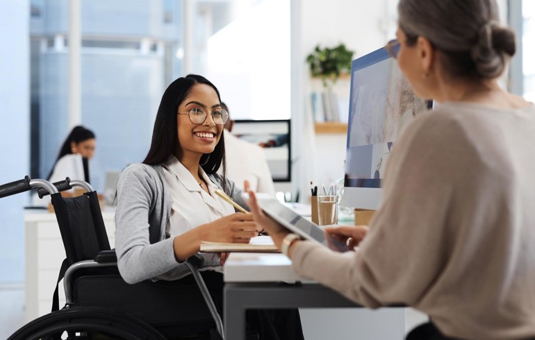 A woman in a wheelchair having a pleasant conversation with another woman while sat at her desk