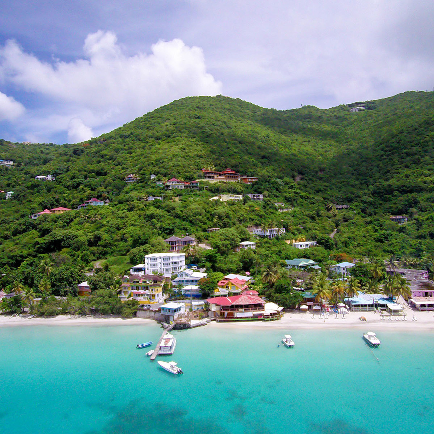 An image of the crystal blue sea with a few boats docked. Green hills in the background with many houses hiding in the shrubbery